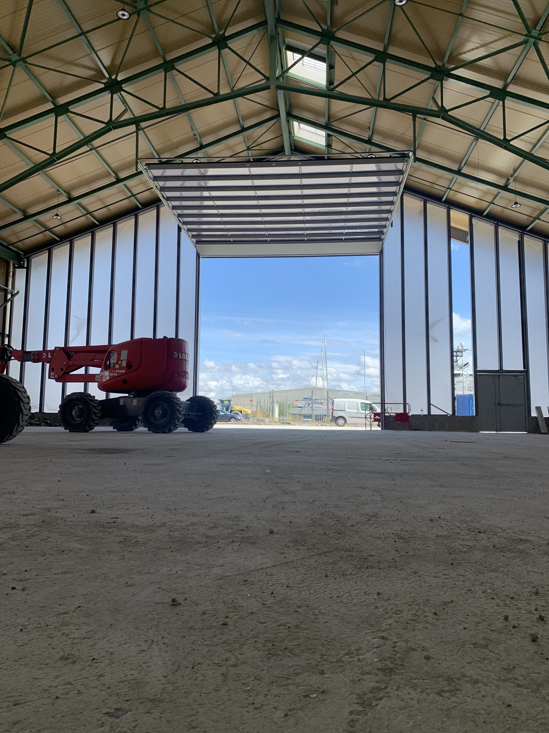 Intérieur d'un grand hangar avec un ascenseur rouge sur la gauche et des portes ouvertes donnant sur un ciel lumineux.