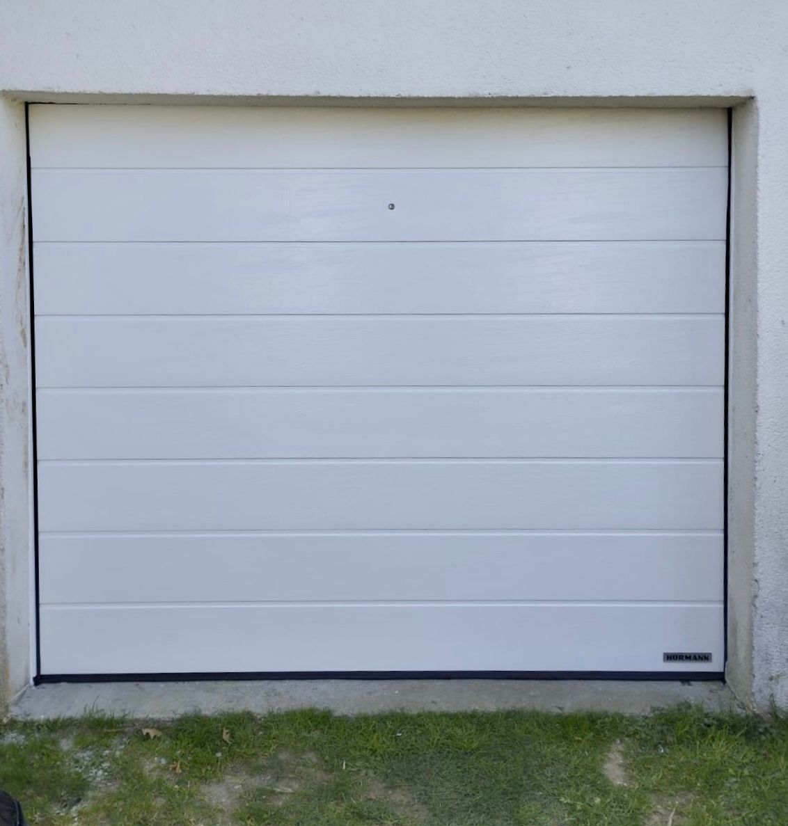 Porte de garage blanche encastrée dans un mur en béton, avec une bande de gazon en dessous.
