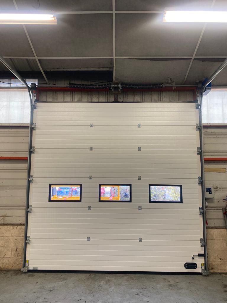 Porte de garage industrielle avec trois petites fenêtres affichant des écrans colorés dans un intérieur ressemblant à un entrepôt.