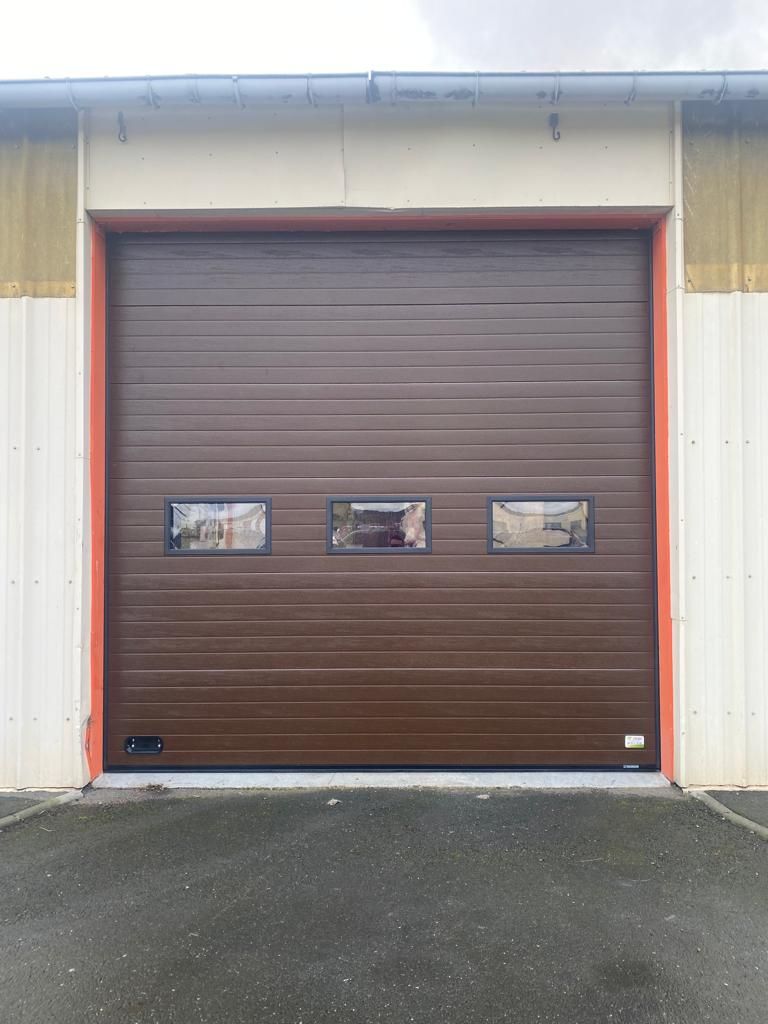 Porte de garage marron avec trois petites fenêtres sur un bâtiment industriel léger