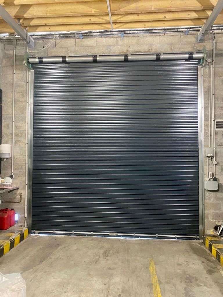 Porte de garage sectionnelle en métal noir fermée dans un espace industriel avec sol en béton et murs en pierre