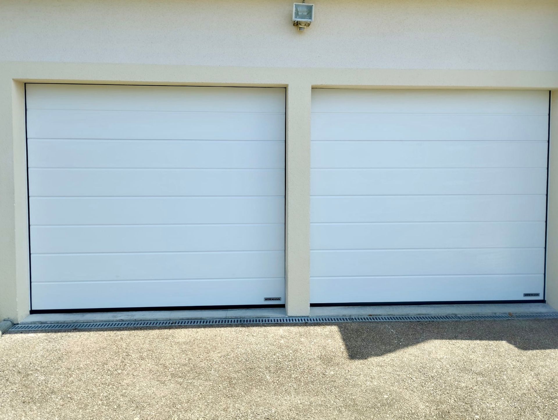 Deux portes de garage blanches encastrées dans un mur beige au-dessus d'une allée de gravier.