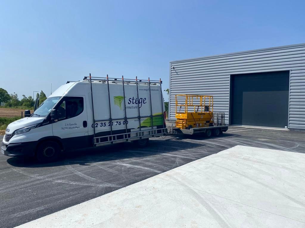 Une camionnette de livraison blanche est garée près d'un quai de chargement d'entrepôt par une journée ensoleillée, avec des palettes de marchandises jaunes à proximité.