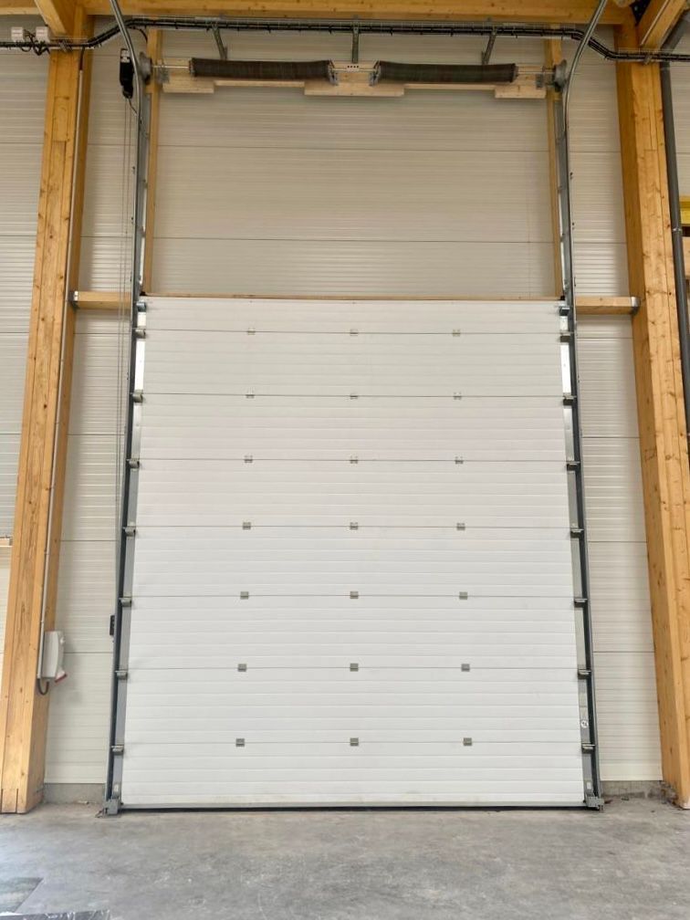 Porte de garage industrielle blanche enroulable fermée, encadrée par des poteaux en bois, à l'entrée d'un entrepôt