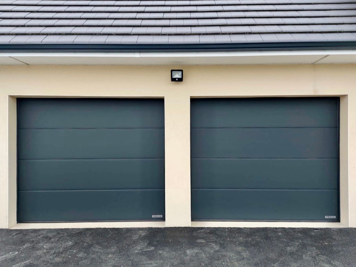 Deux portes de garage gris foncé sur une façade de maison beige, éclairées par une petite applique murale.