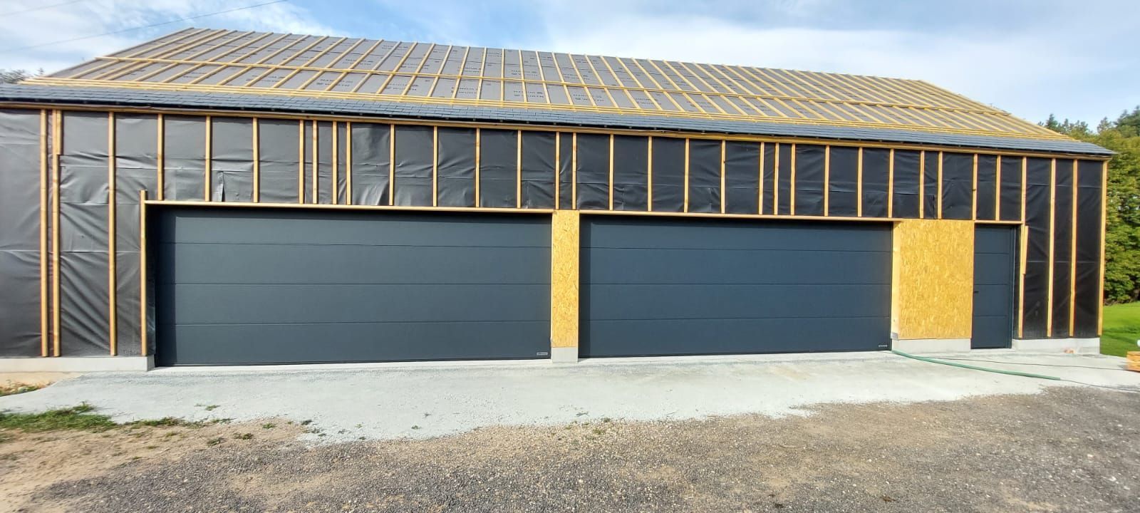 Extérieur de maison inachevé avec charpente apparente, portes de garage sombres et revêtement en contreplaqué sous un toit de tuiles.