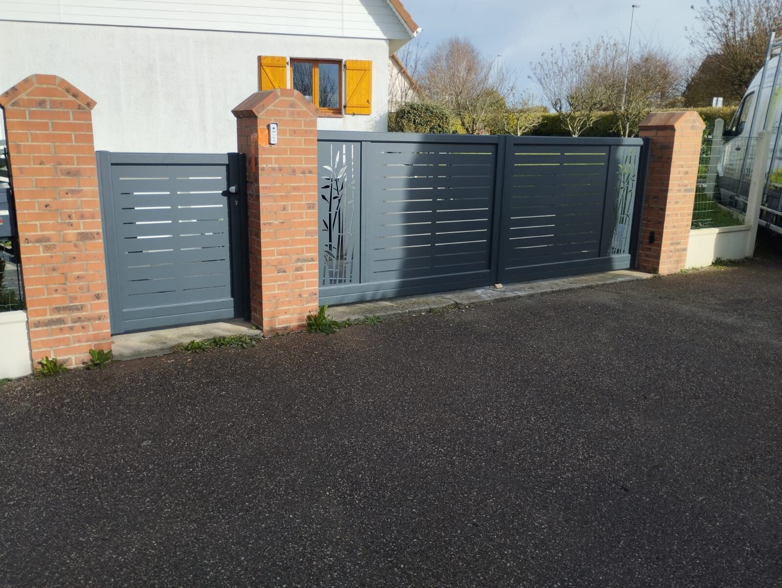 Portail d'allée gris entre des piliers de briques devant une maison blanche et une rue pavée
