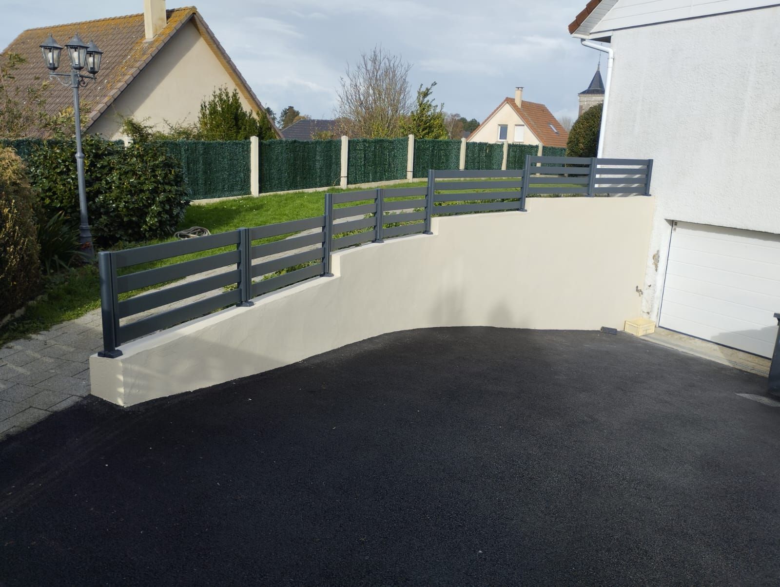 Mur de soutènement incurvé blanc avec des balustrades gris foncé, bordant une allée pavée dans un quartier résidentiel.