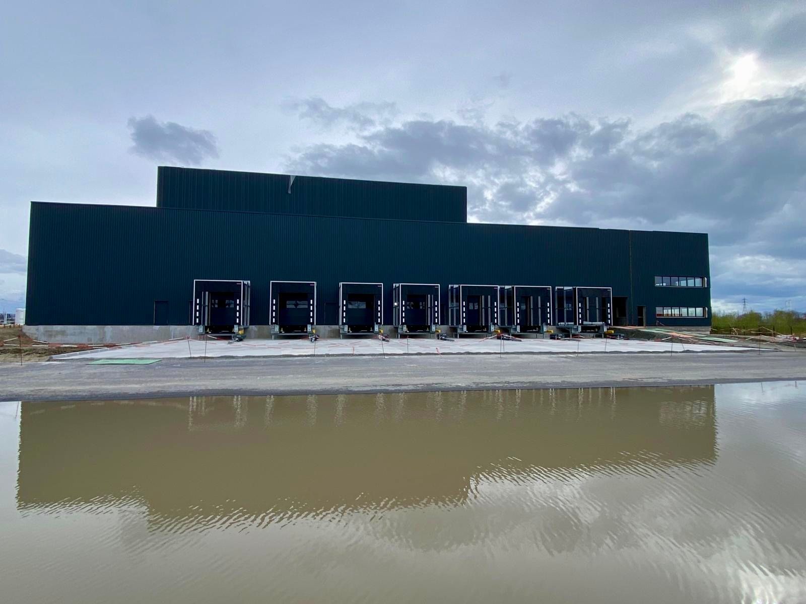 Bâtiment industriel vert foncé avec quais de chargement à côté d'une flaque boueuse sous un ciel nuageux