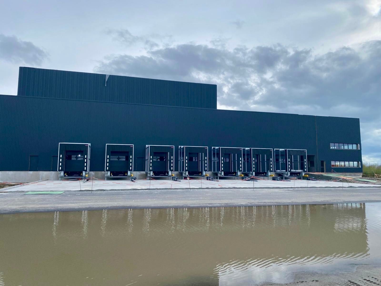 Grand entrepôt bleu foncé avec quais de chargement à côté d'une zone inondée et boueuse sous un ciel nuageux.