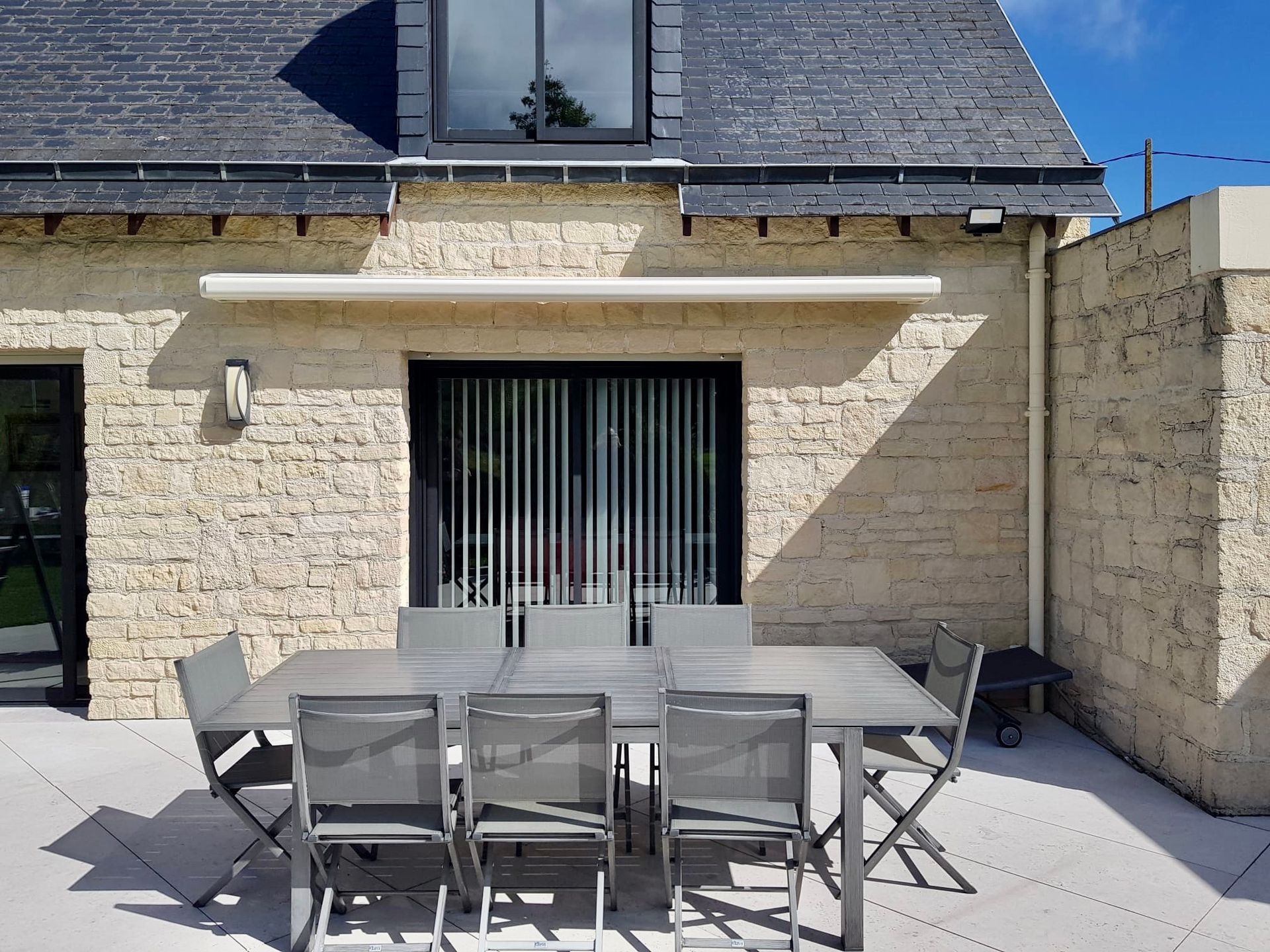 Terrasse en pierre avec table et chaises de jardin devant une maison dotée de grandes portes coulissantes