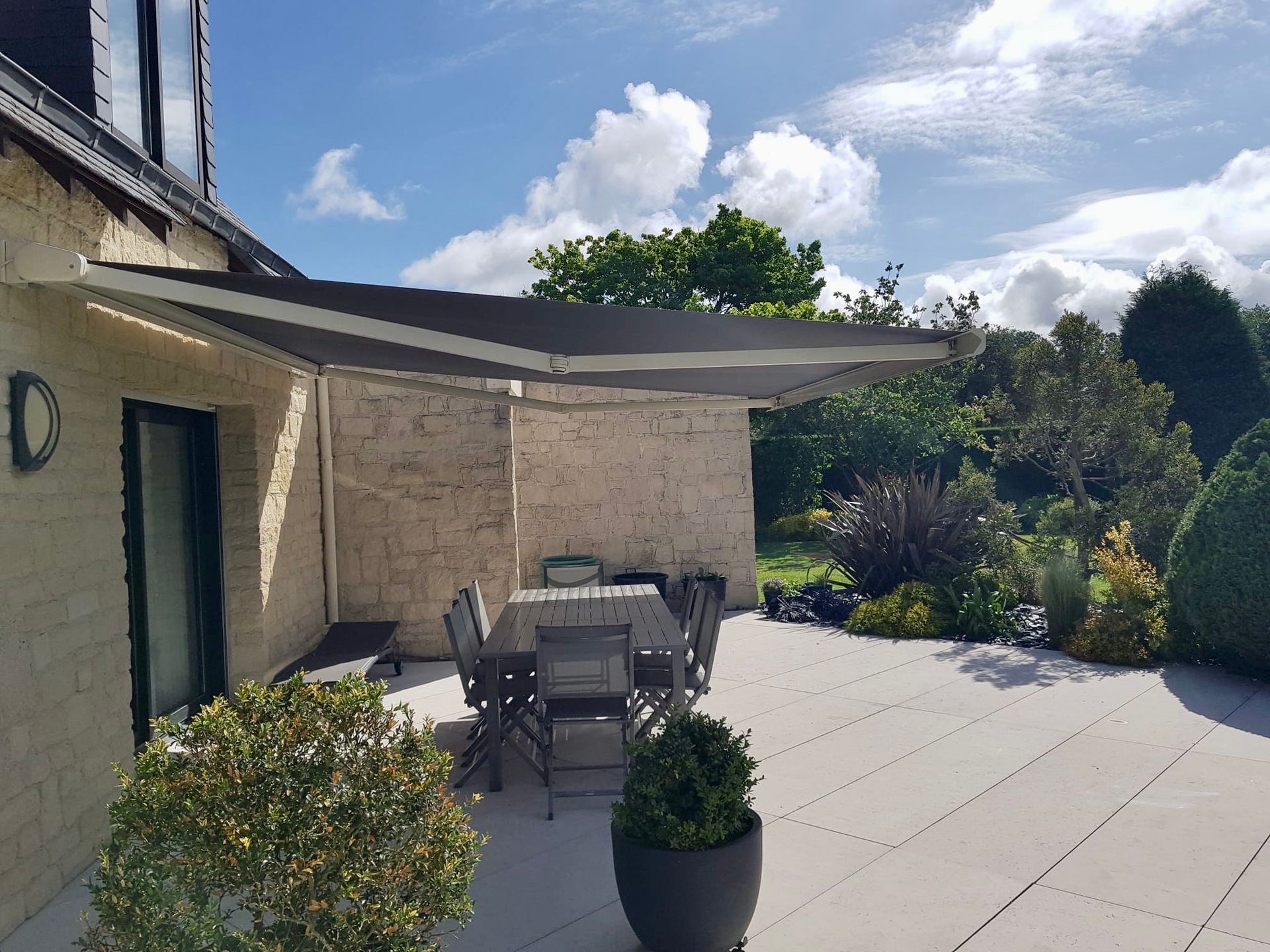 Terrasse en pierre avec table à manger extérieure, plantes en pot et voile d'ombrage à côté d'une maison sous un ciel ensoleillé