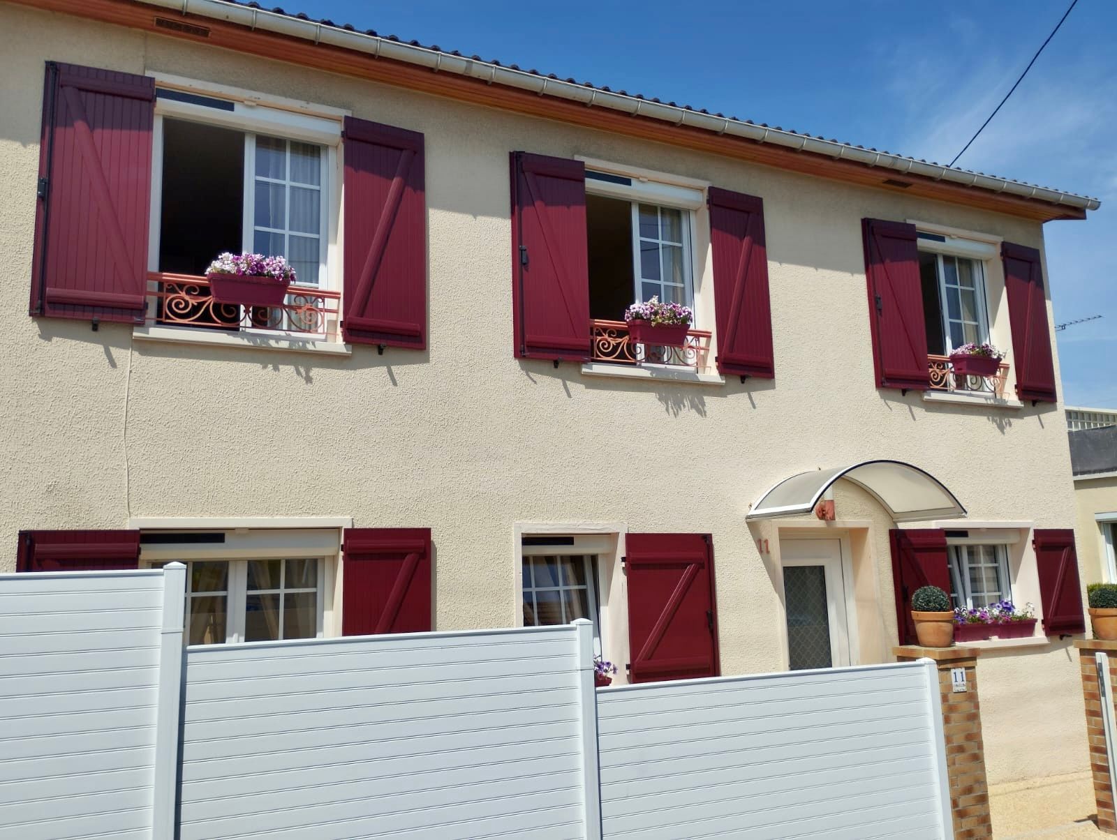 Maison beige à deux étages avec volets bordeaux et clôture blanche sous un ciel bleu