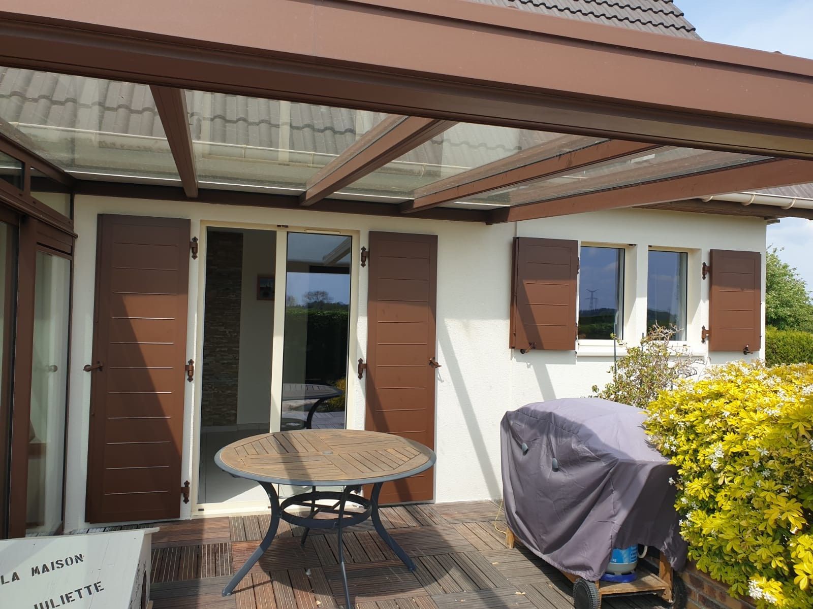 Terrasse avec table ronde, barbecue couvert et maison blanche aux volets marron sous un auvent.