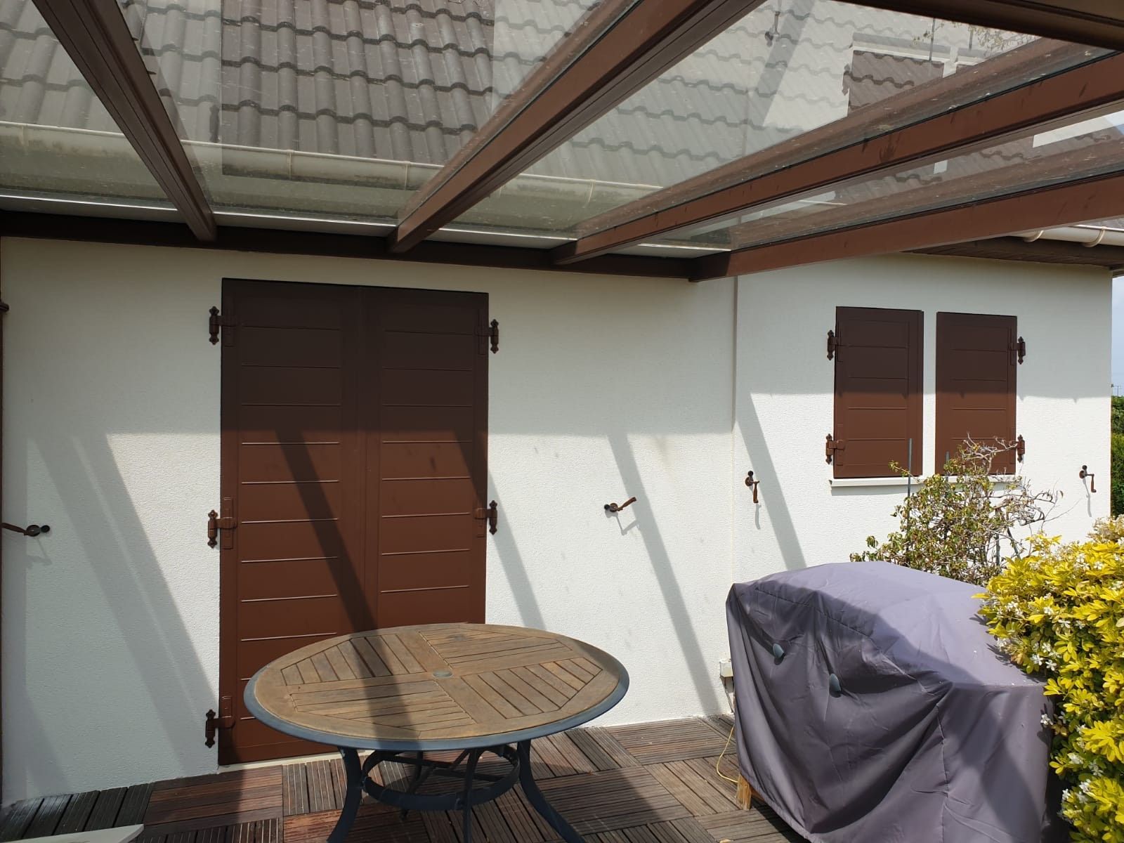 Terrasse avec table ronde, barbecue couvert, porte marron et fenêtre à volets sous verrière