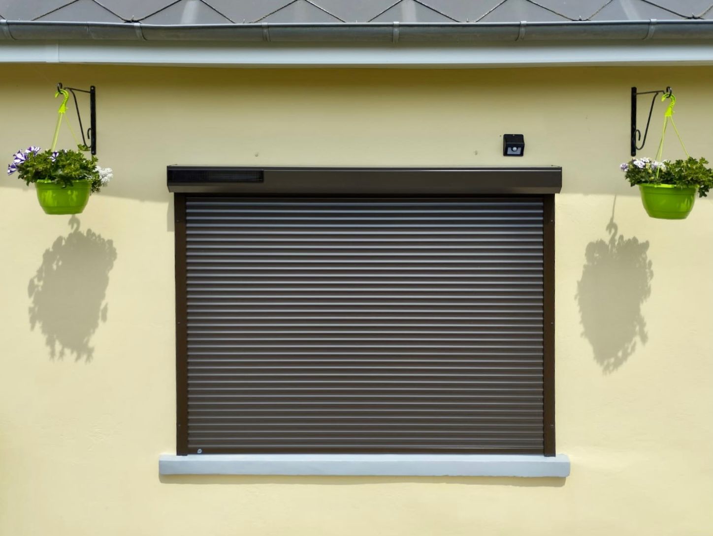Volet de fenêtre marron fermé sur un mur beige avec des pots de fleurs verts suspendus de chaque côté