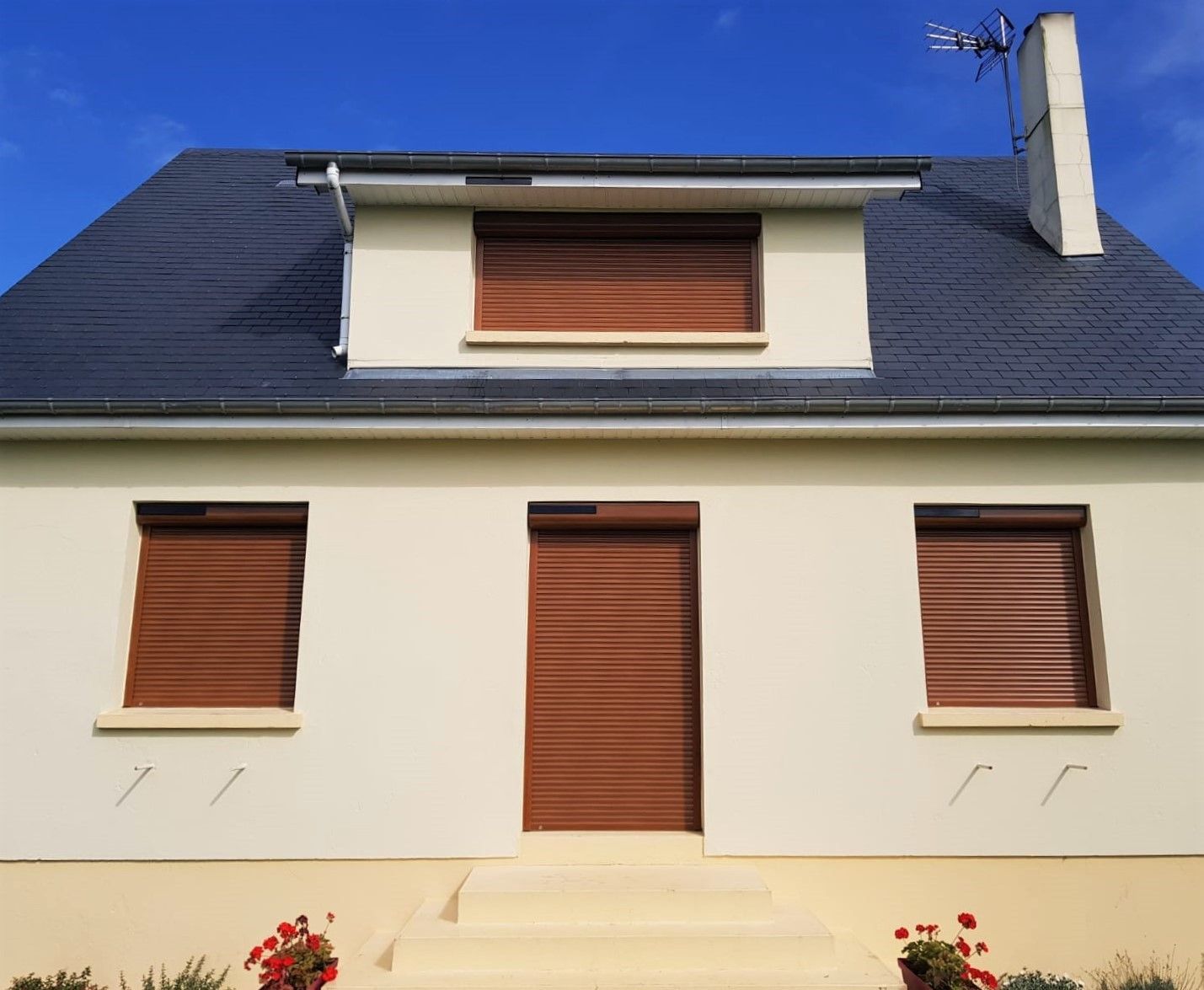Maison couleur crème au toit foncé et aux volets marron fermés, sous un ciel bleu.