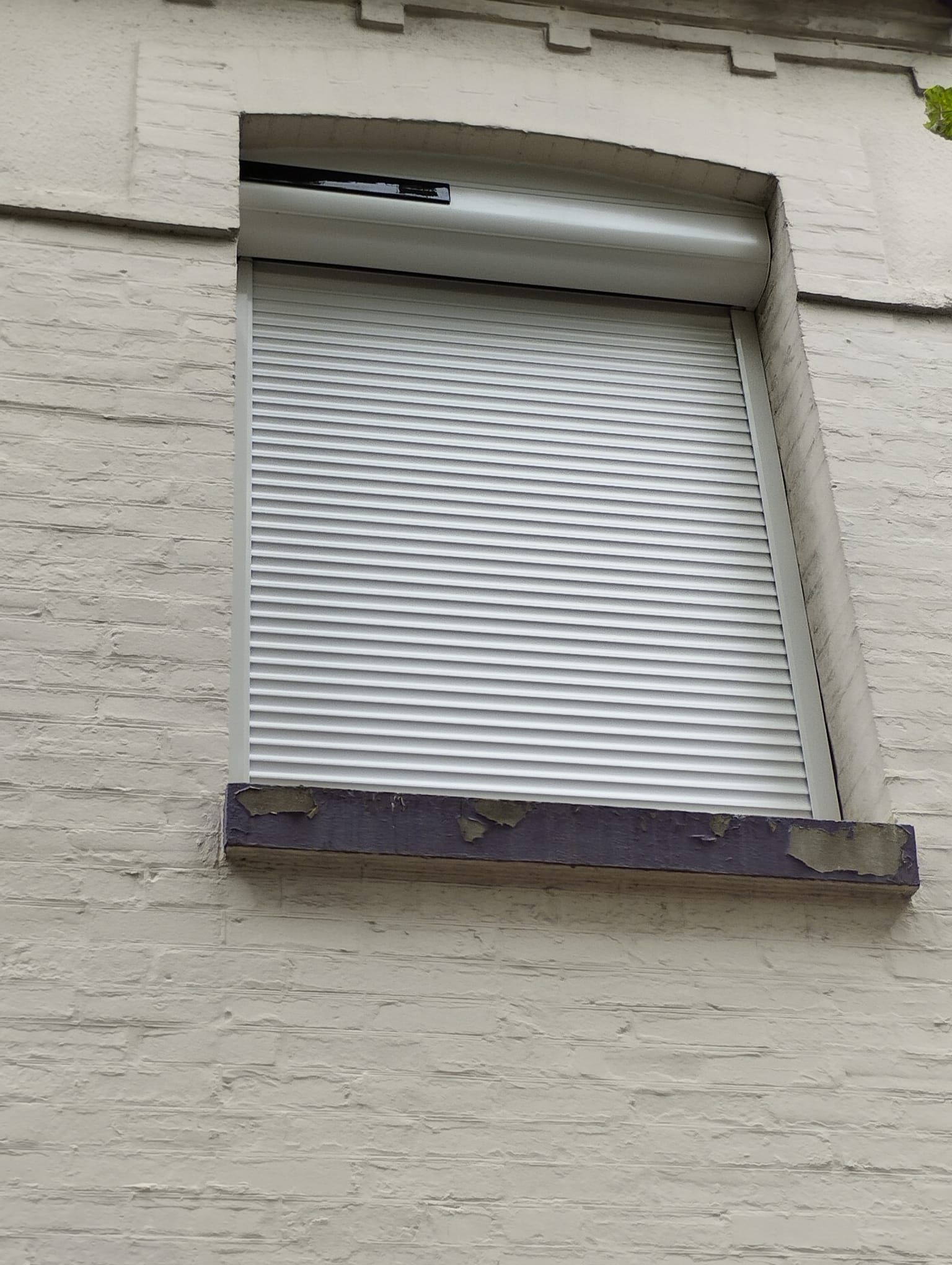 Fenêtre à volets blancs fermés sur un mur de briques beiges avec un appui de fenêtre inférieur endommagé