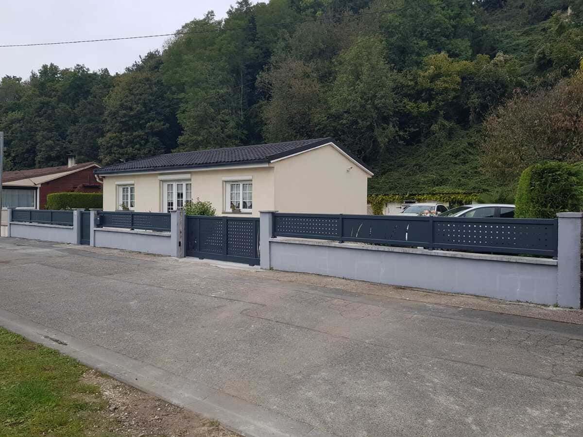 Petite maison couleur crème avec une clôture sombre, située sur un chemin de gravier à côté d'une colline boisée.
