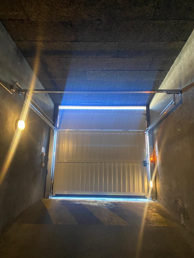 Un tunnel en béton sombre mène à un portail métallique ondulé fermé, éclairé par des lumières bleues et chaudes.