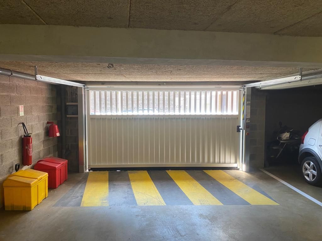 Parking souterrain avec un portail blanc fermé, des bandes jaunes au sol et une voiture garée sur la droite.