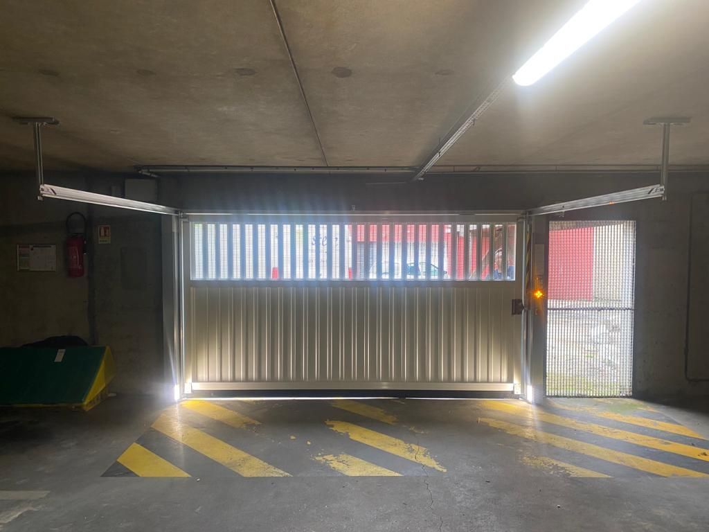Entrée du parking souterrain avec portail métallique fermé, marquages ​​de signalisation rayés et porte latérale éclairée par une lumière vive.