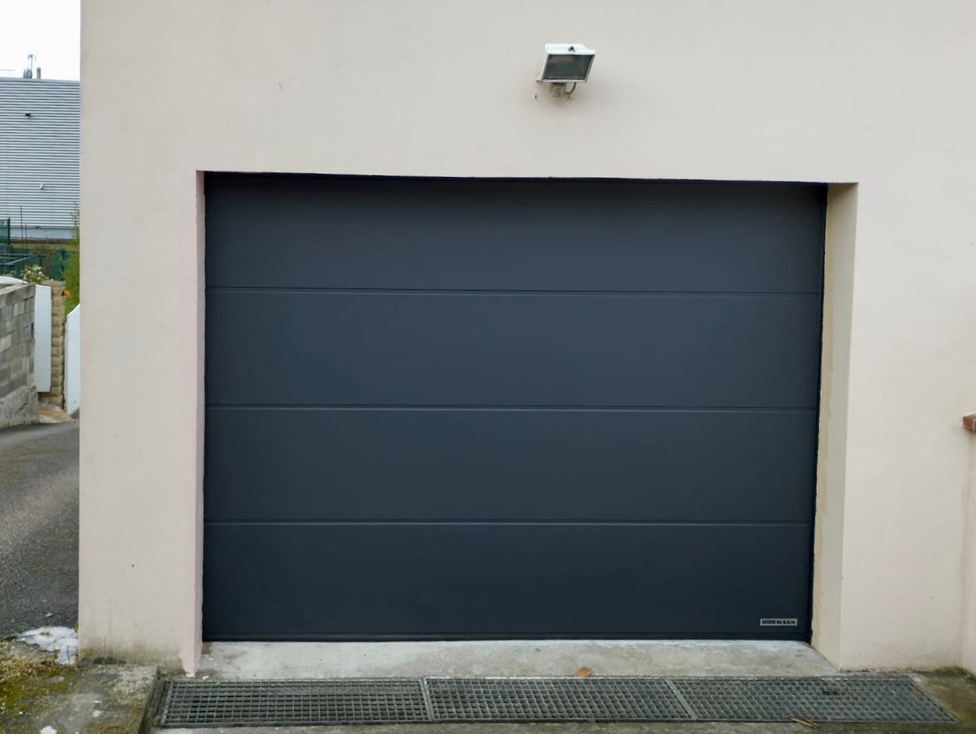 Porte de garage gris foncé sur un bâtiment beige, avec un petit éclairage au-dessus et une grille d'évacuation en dessous.