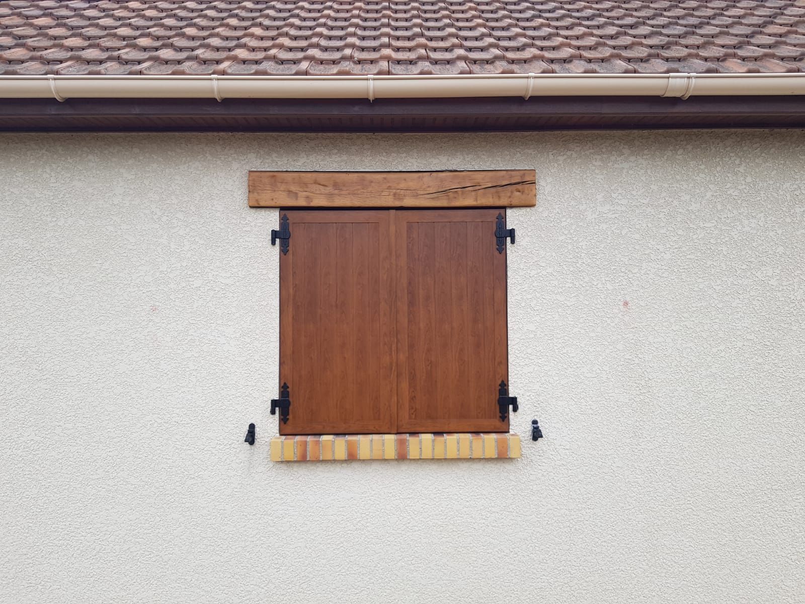 Volet roulant marron fermé sur un mur beige sous un toit de tuiles.