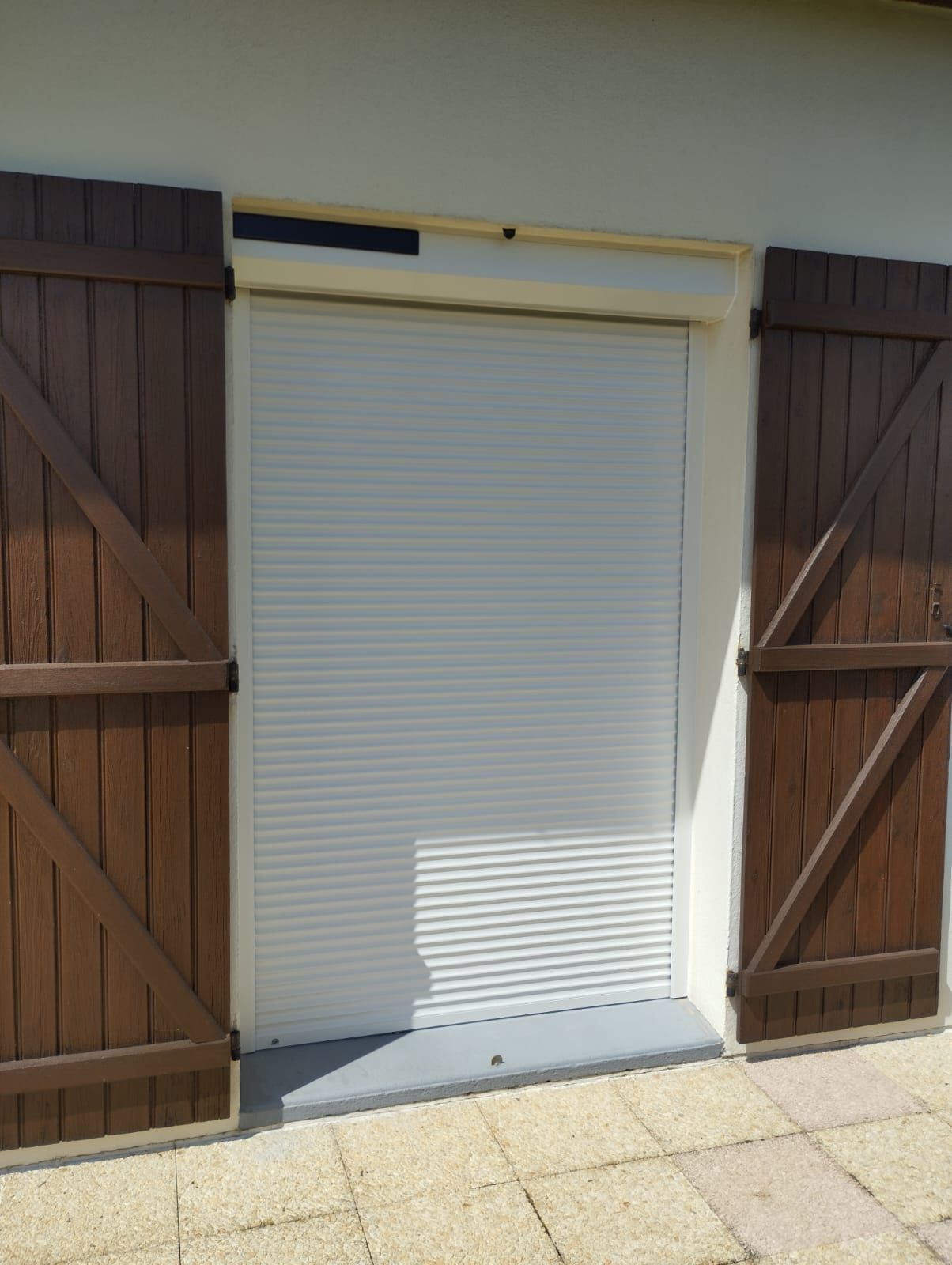 Volet roulant blanc fermé entre des portes en bois brun sur une terrasse ensoleillée