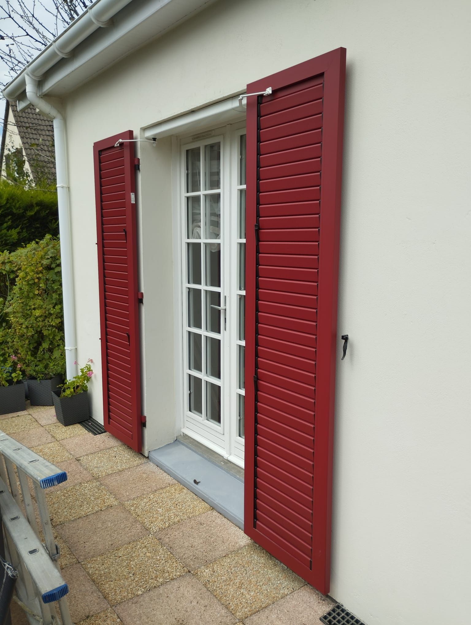 Portes-fenêtres blanches à volets rouges donnant sur une terrasse pavée.