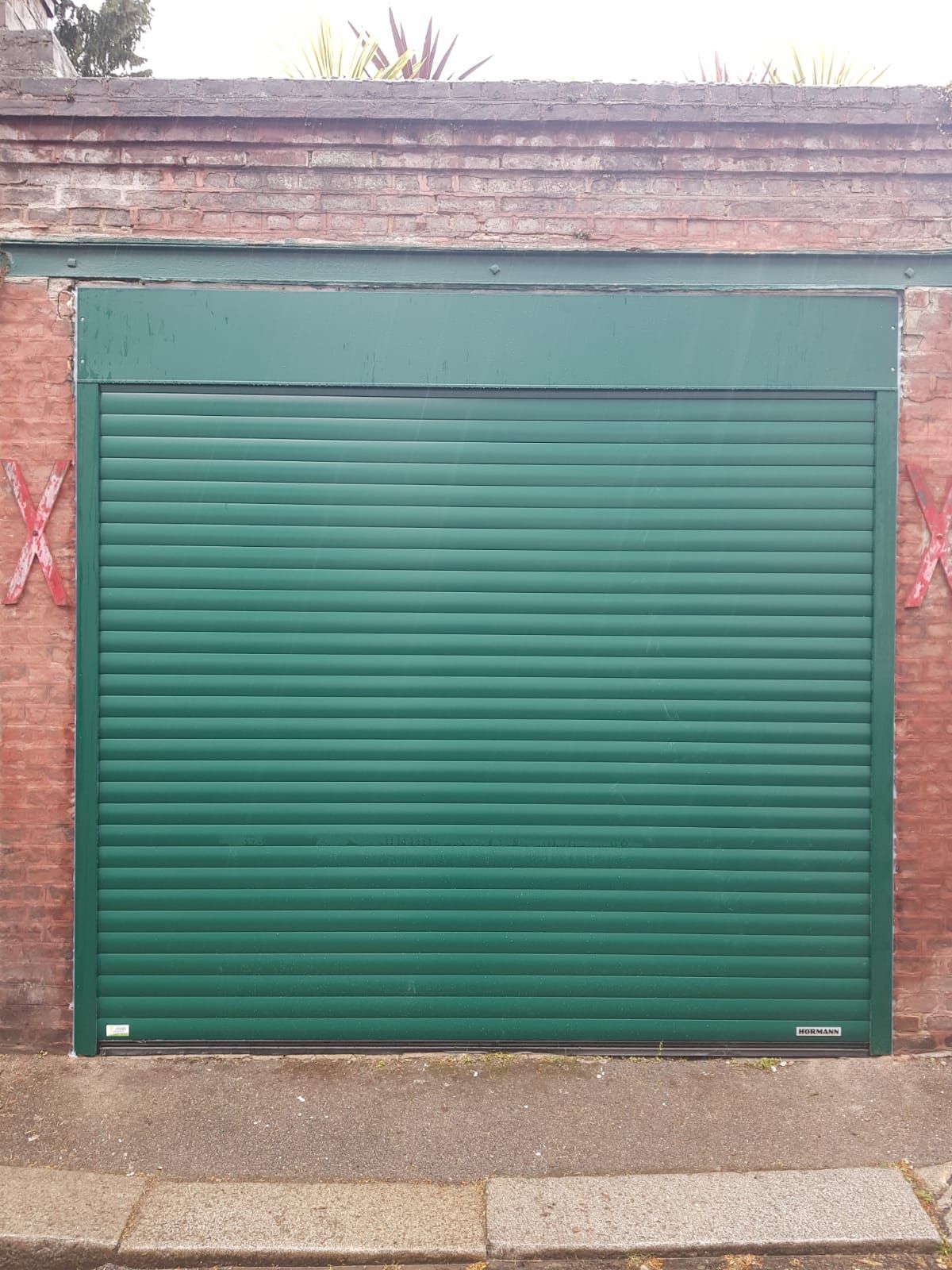 Porte de garage en métal vert encastrée dans un mur de briques, avec un trottoir en béton devant.