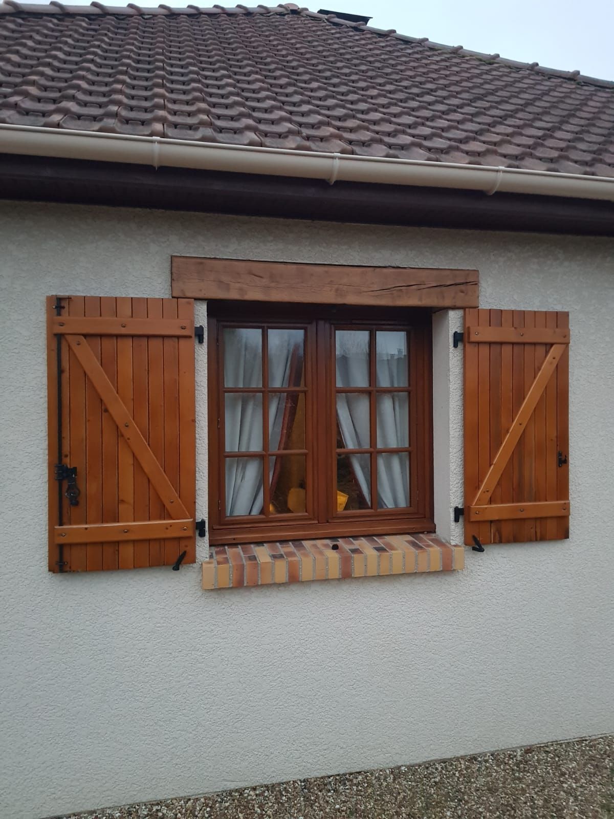Fenêtre de maison avec volets en bois brun et toit de tuiles au-dessus.