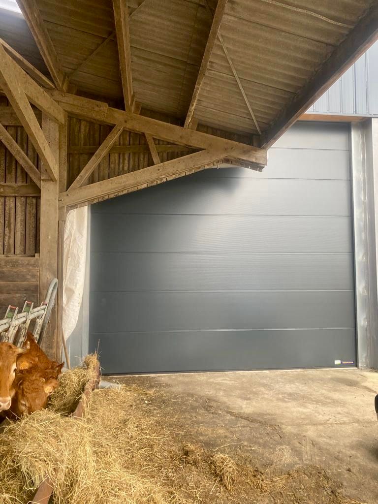 Des poules picorent de la paille près d'une grande porte de grange grise, sous un toit en bois.