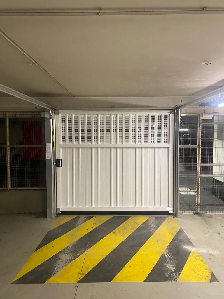 Portail métallique blanc dans un parking souterrain, au-dessus de bandes de signalisation jaunes et noires.