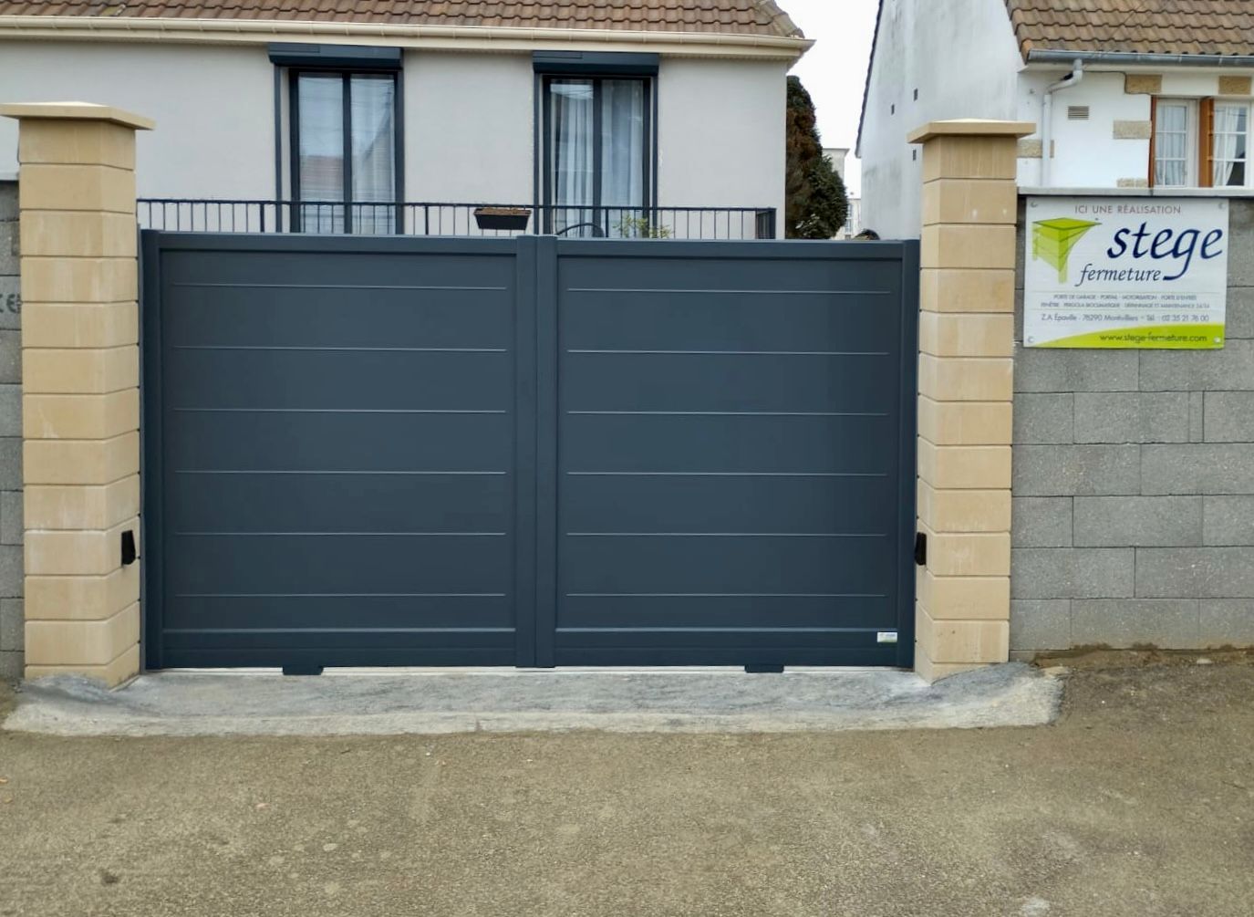Portail d'allée en métal gris foncé entre des piliers en pierre, devant une maison avec un panneau « À vendre » sur la droite.