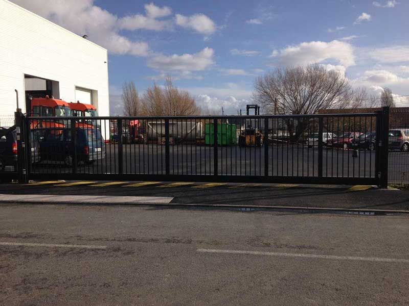 Portail coulissant en métal noir barrant un parking à côté d'un bâtiment blanc sous un ciel partiellement nuageux