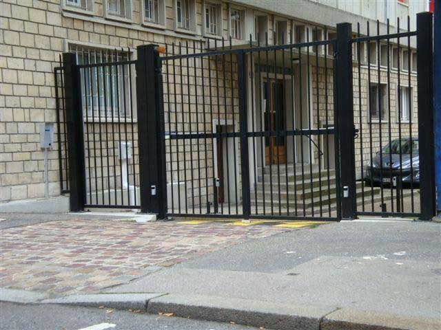 Un portail et une clôture en métal noir bloquent un trottoir à côté d'un bâtiment en pierre et d'une voiture garée.