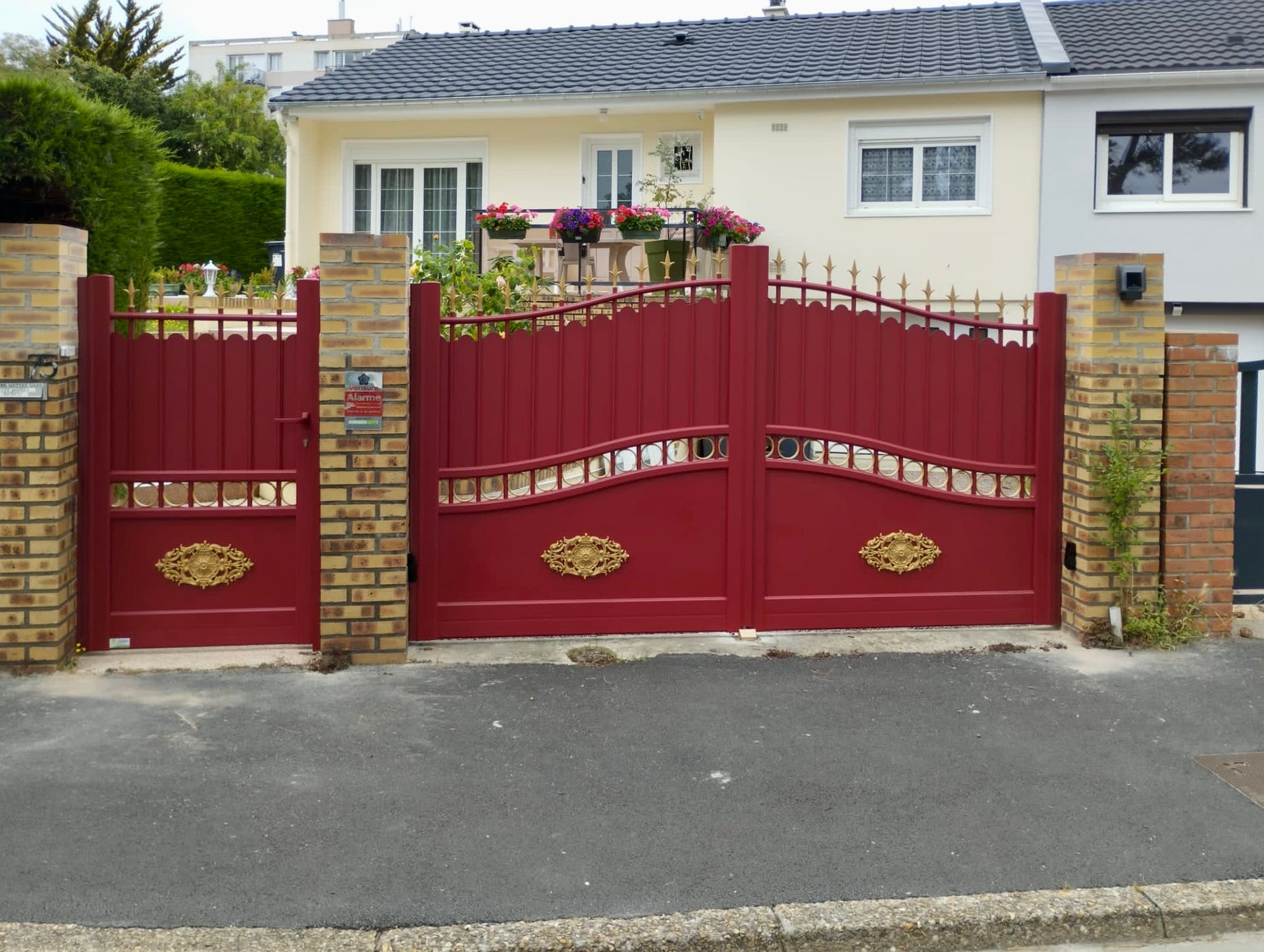 Portail double rouge avec piliers en briques devant une maison de plain-pied et un jardin