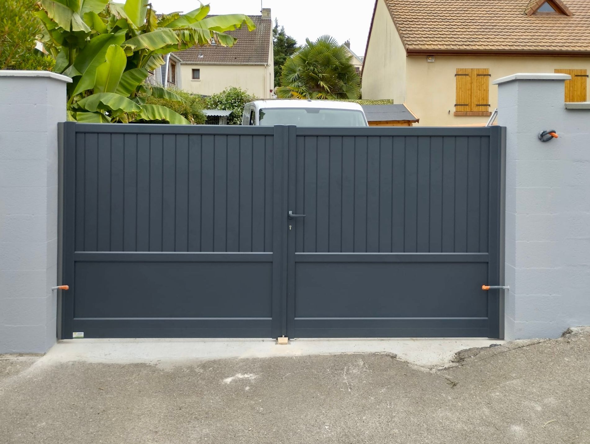 Un portail double gris foncé est encastré dans un mur gris clair, laissant apparaître une maison et de la verdure derrière.