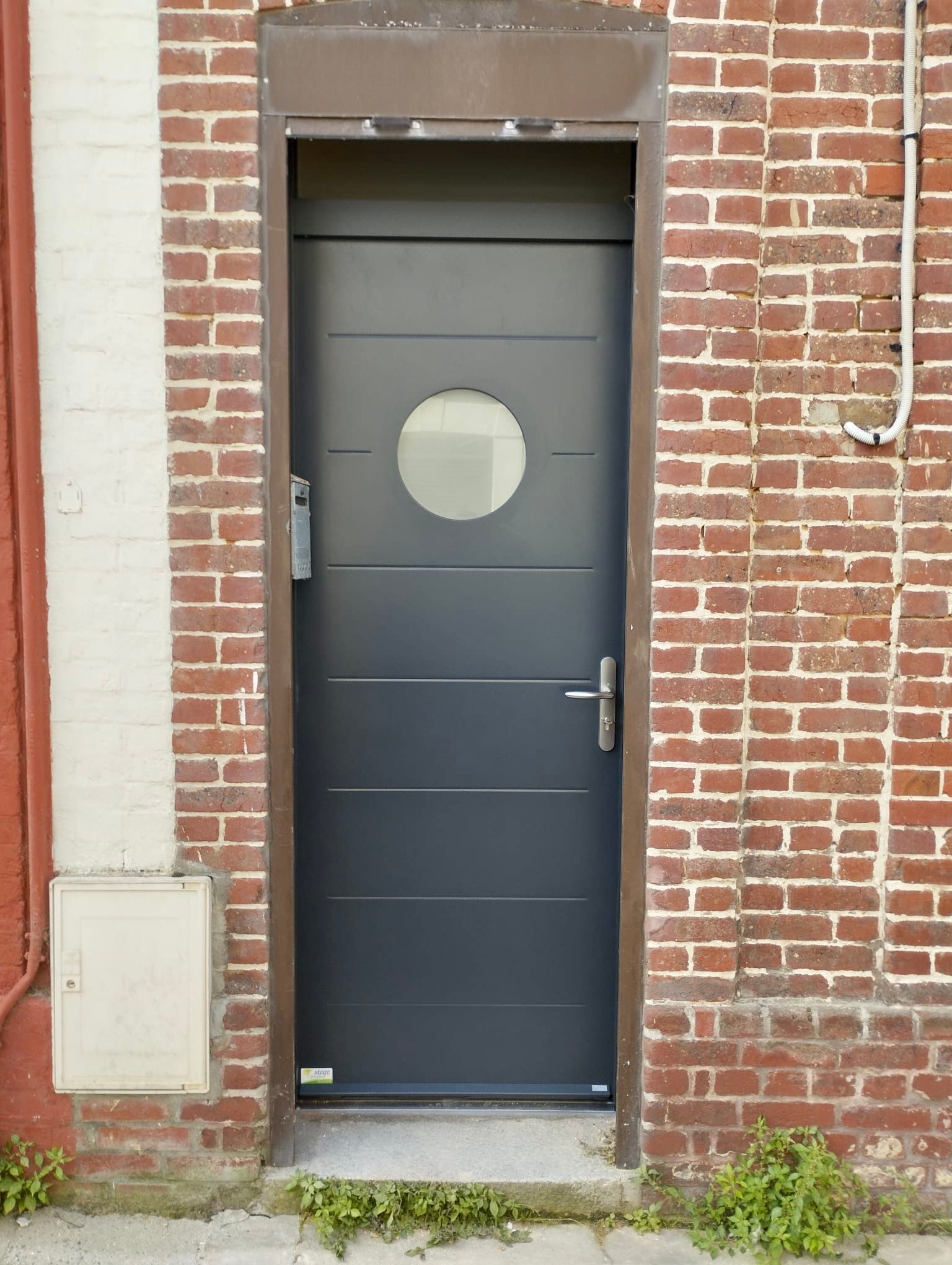 Porte d'entrée gris foncé avec fenêtre ronde encastrée dans un mur de briques rouges