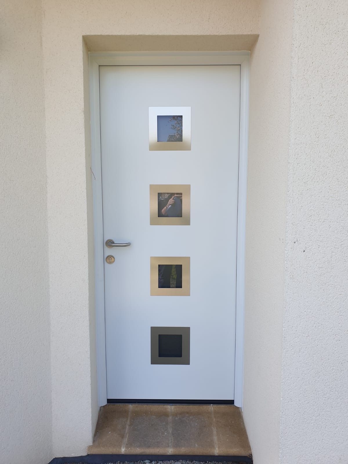 Porte d'entrée blanche avec quatre petites fenêtres carrées, encastrée dans un mur beige avec une marche carrelée.