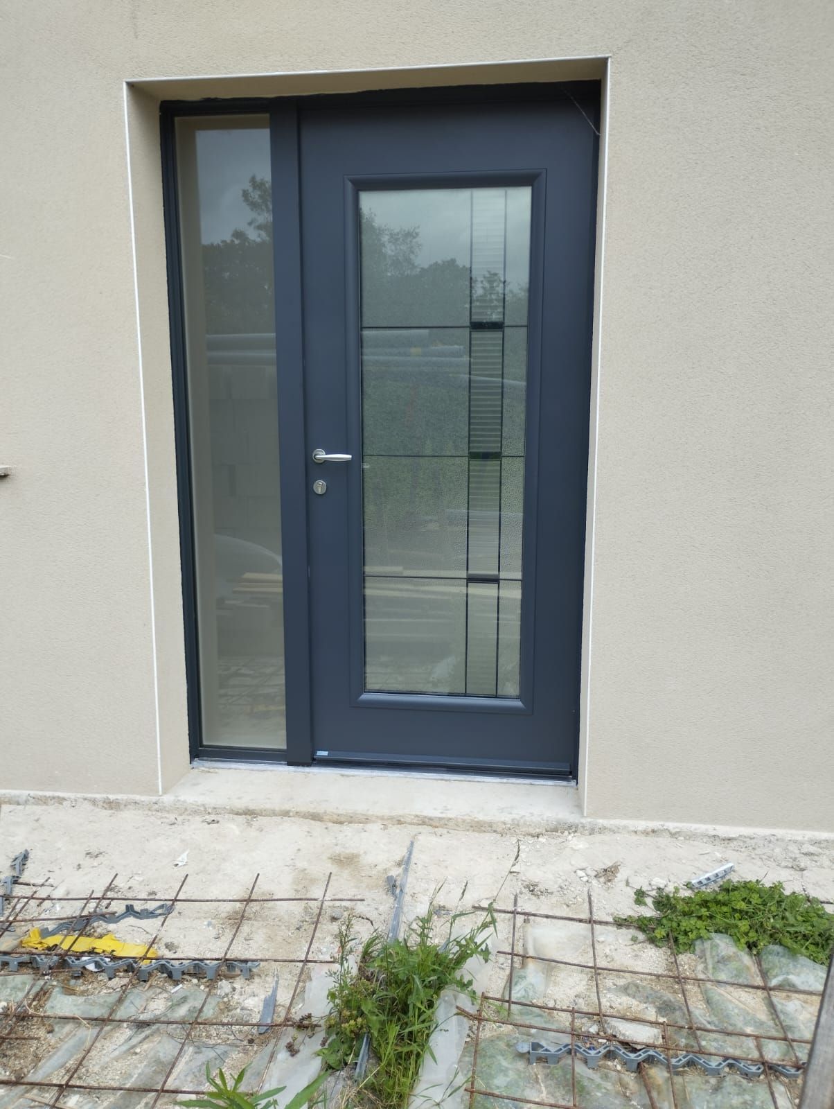 Porte d'entrée bleu foncé avec panneaux de verre dépoli encastrée dans un mur extérieur beige, avec un chemin carrelé et des mauvaises herbes en contrebas.