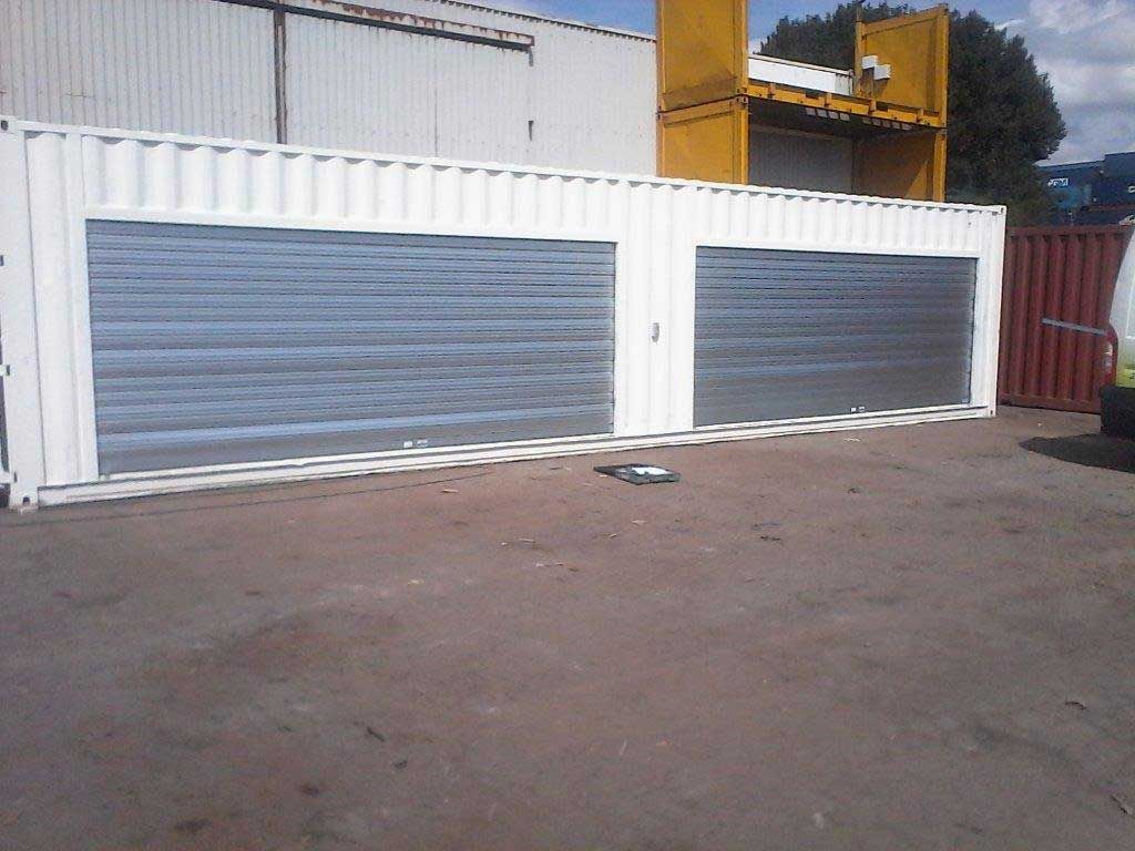 Deux portes de garage enroulables grises sur un bâtiment industriel blanc à côté d'un terrain pavé