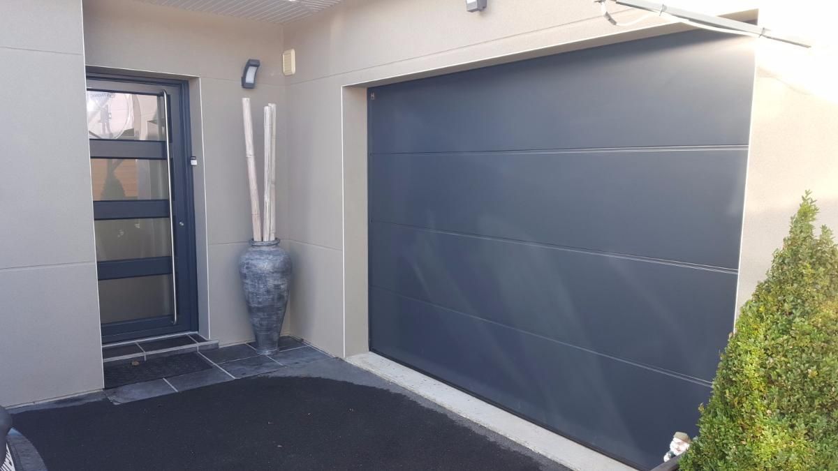 Entrée de maison moderne avec une porte de garage gris foncé, une porte d'entrée noire et une plante en pot.