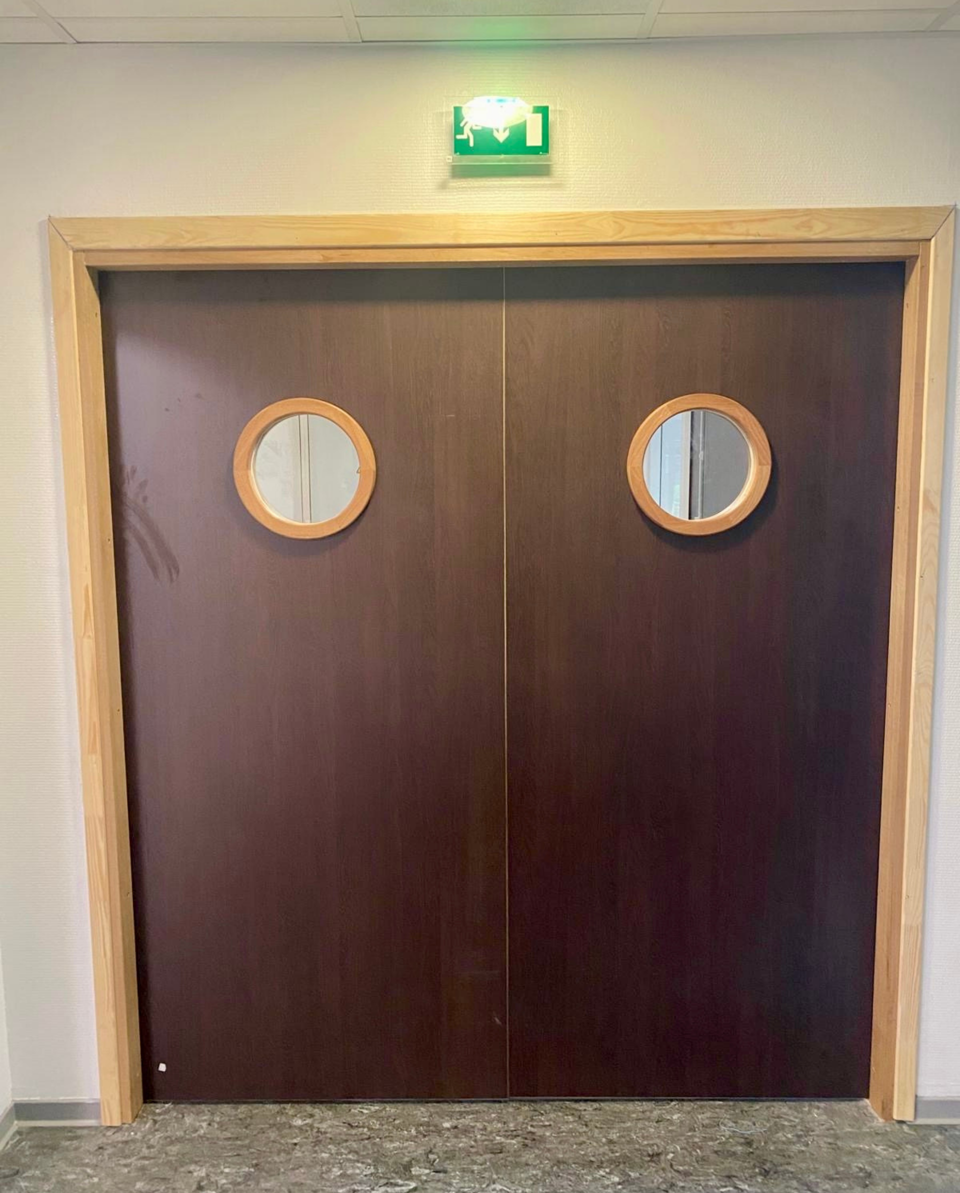 Double porte en bois avec fenêtres rondes dans un couloir, surmontée d'un panneau de sortie vert.