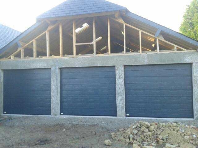 Trois portes de garage sombres sur un bâtiment en béton en construction, avec une charpente de toit apparente.