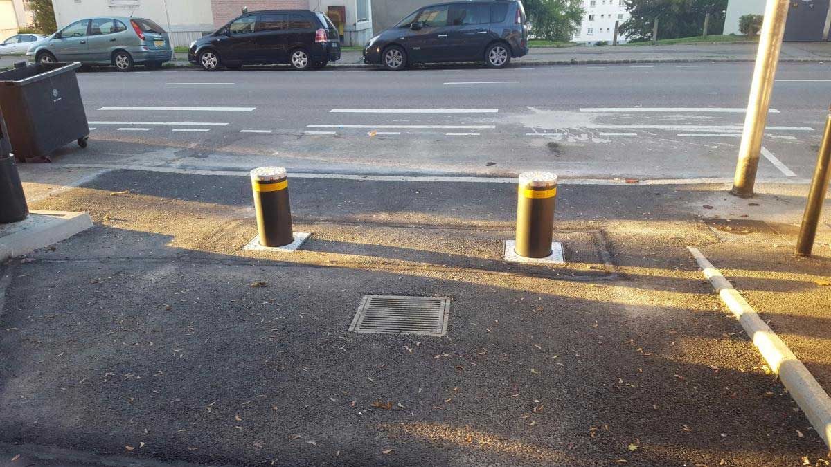 Sortie de parking avec deux bornes jaunes et une route au-delà, vue en plein soleil.