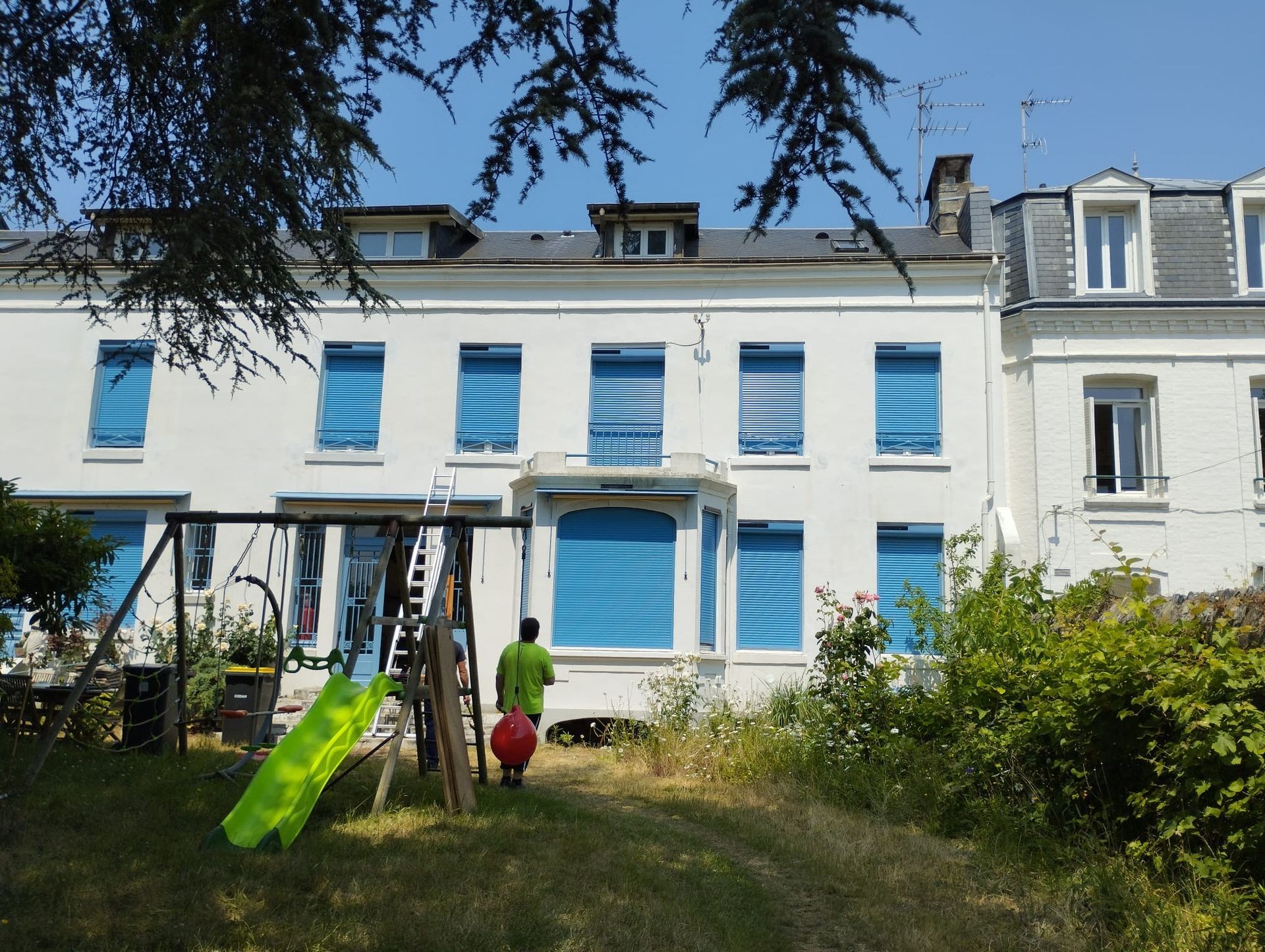 Maison de ville blanche avec des volets bleus et un jardin, avec un enfant qui joue près d'un toboggan vert devant.