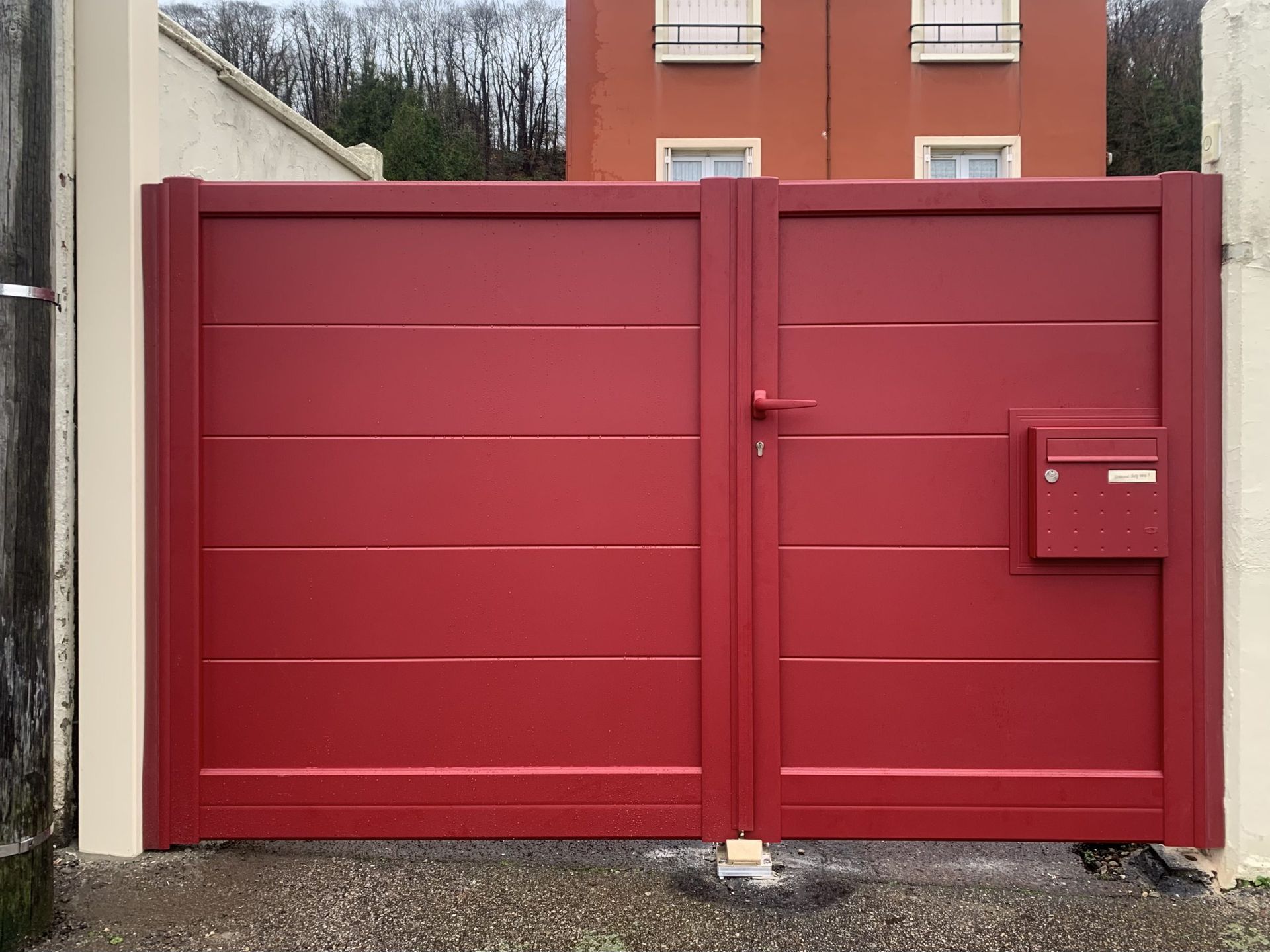 Portail double en métal rouge avec une petite boîte aux lettres, installé dans une allée de gravier devant un bâtiment.