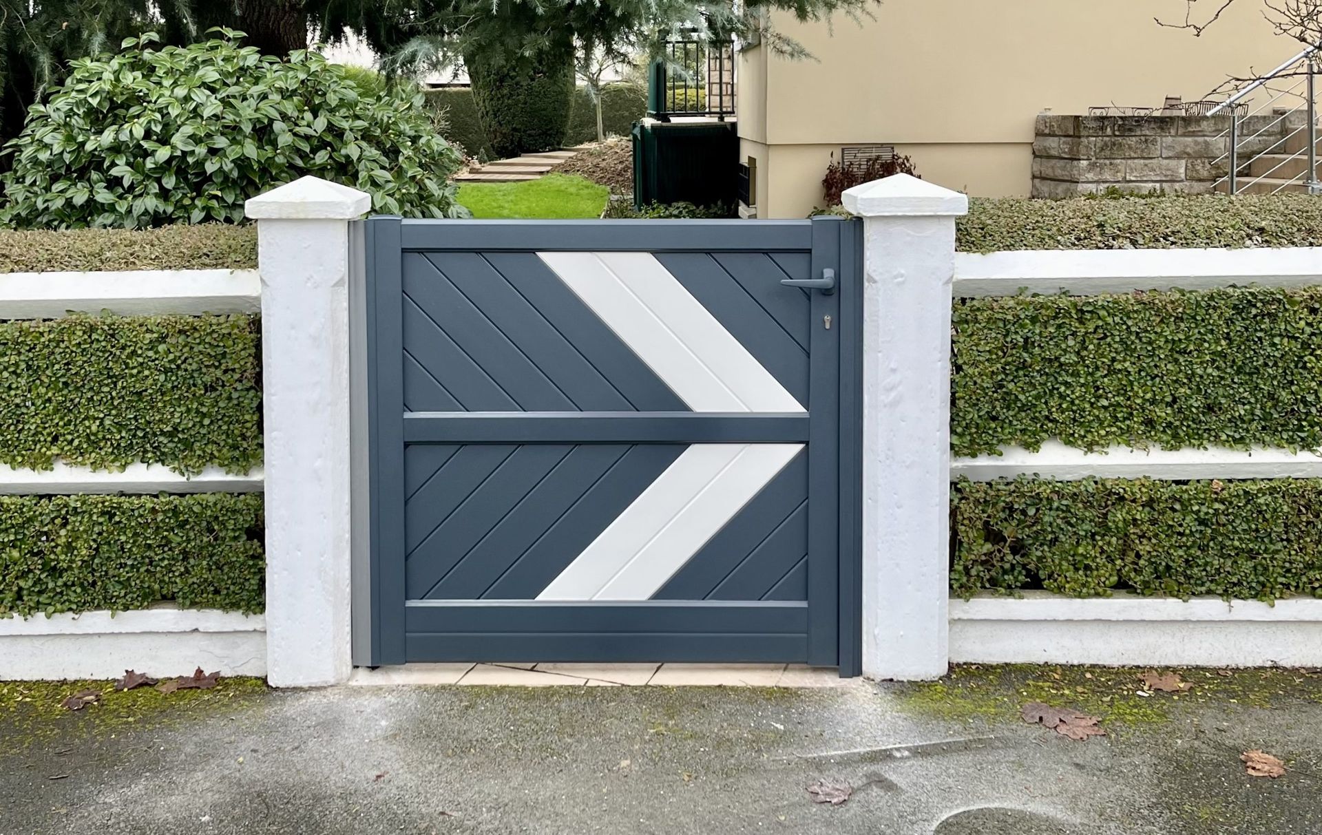 Portail de jardin gris foncé avec panneaux à chevrons blancs entre des poteaux de clôture blancs et des haies.